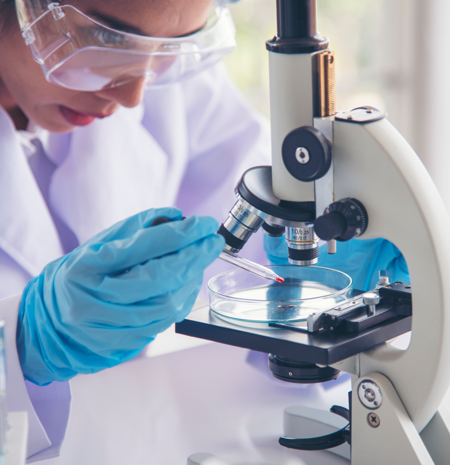 A person in lab coat and goggles looking at an object through a microscope.